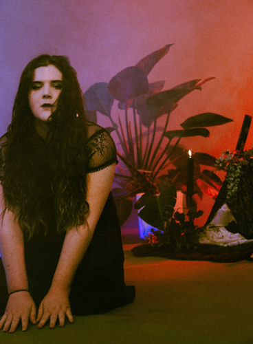 Babydoll, wearing a black dress with lace accents, kneeling in a room with red and blue lighting and several plants behind her.