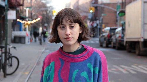 Frankie Cosmos (Greta Kline) standing on the street in a funky pink, blue, and teal sweater with a perfect bob haircut.