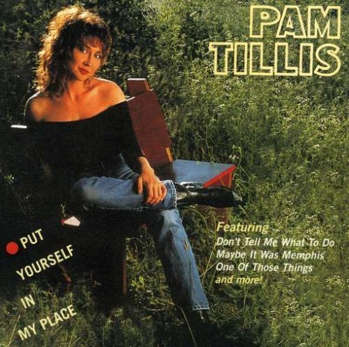 Pam Tillis on a bench in a grassy field, wearing a black off the shoulder top, with denim jeans and black leather boots.